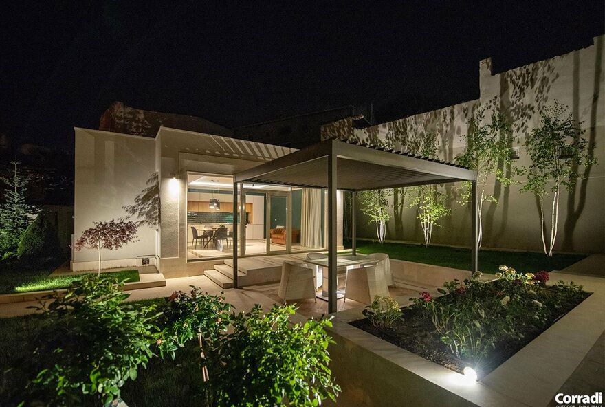 Ein beleuchteter Garten bei Nacht mit einer von einem Lamellendach geschützten Terrasse