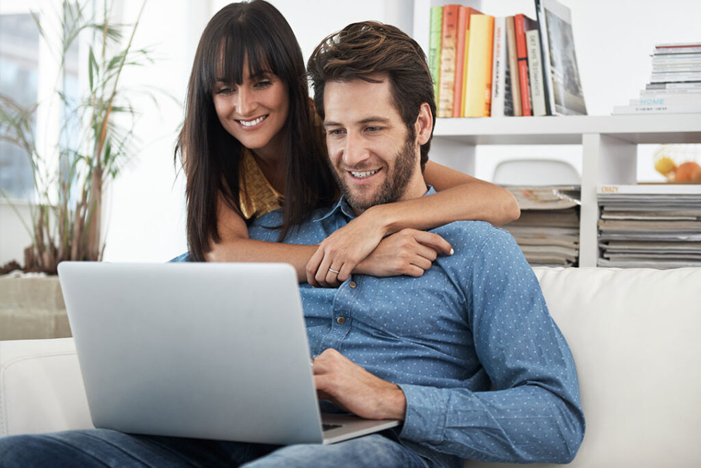 Eine Frau umarmt einen Mann von hinten, während er einen Laptop benutzt. Sie sitzen auf einem weißen Sofa in einem hellen Raum. Im Hintergrund stehen Bücher und eine Pflanze. Beide lächeln und wirken entspannt.