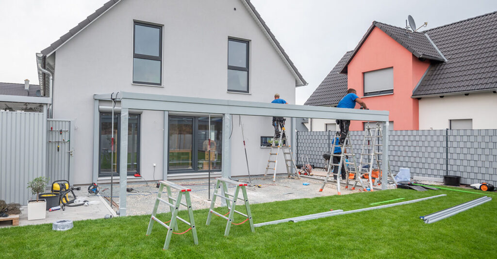 Bauarbeiter errichten an der Rückseite eines zweistöckigen Hauses mit grauen und rosa Außenwänden eine moderne Glasterrassenüberdachung. Der Terrassenbereich ist von gepflegtem grünem Gras umgeben.