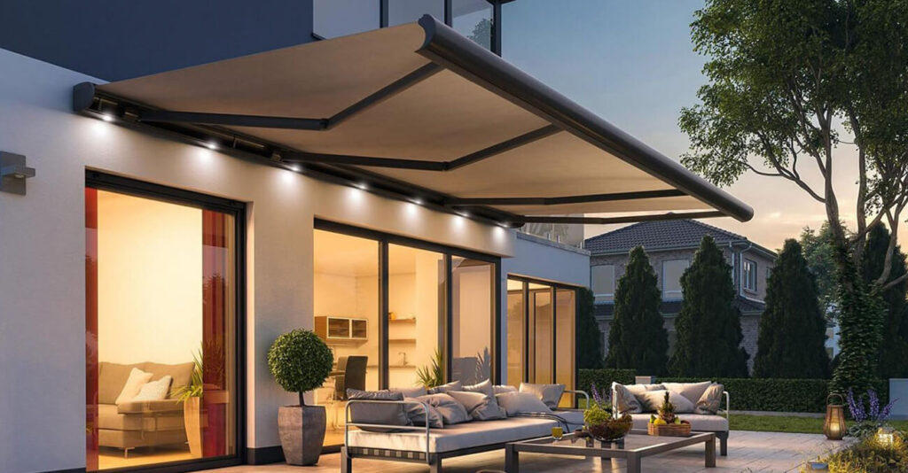 Moderne Terrasse mit einer großen, einziehbaren Markise, die sich über die Sitzgelegenheiten im Freien erstreckt. Der Bereich verfügt über gepolsterte Sofas und einen Glastisch. Das Haus hat große Fenster mit warmer Beleuchtung im Inneren. Üppiges Grün und Bäume säumen den Raum.
