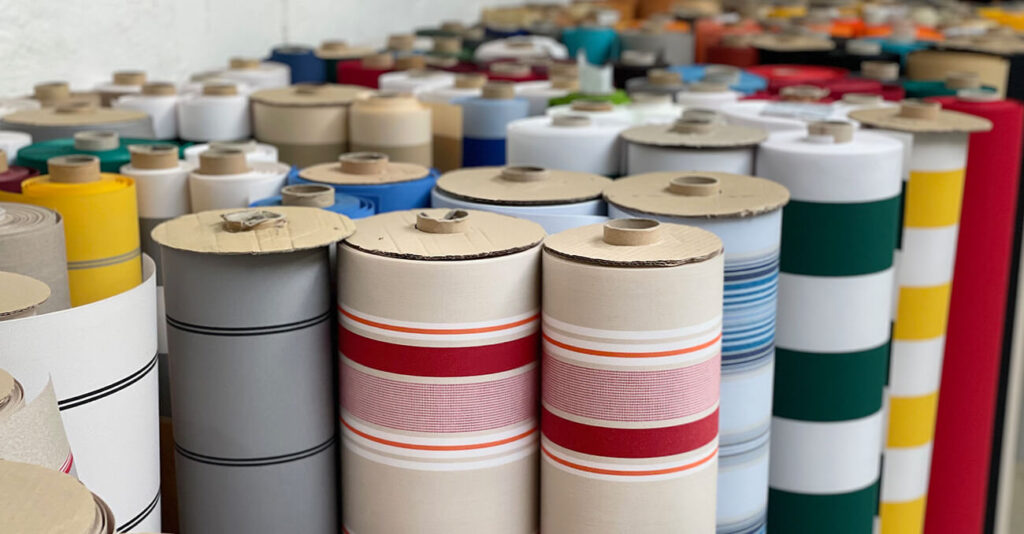 Reihen aufrecht stehender Stoffrollen mit verschiedenen Mustern, darunter Streifen und einfarbige Stoffe. Die Rollen sind dicht gepackt und die Farben reichen von Rot und Grün bis hin zu Gelb, Blau und mehr. Im Hintergrund sind weitere gestapelte Rollen zu sehen.