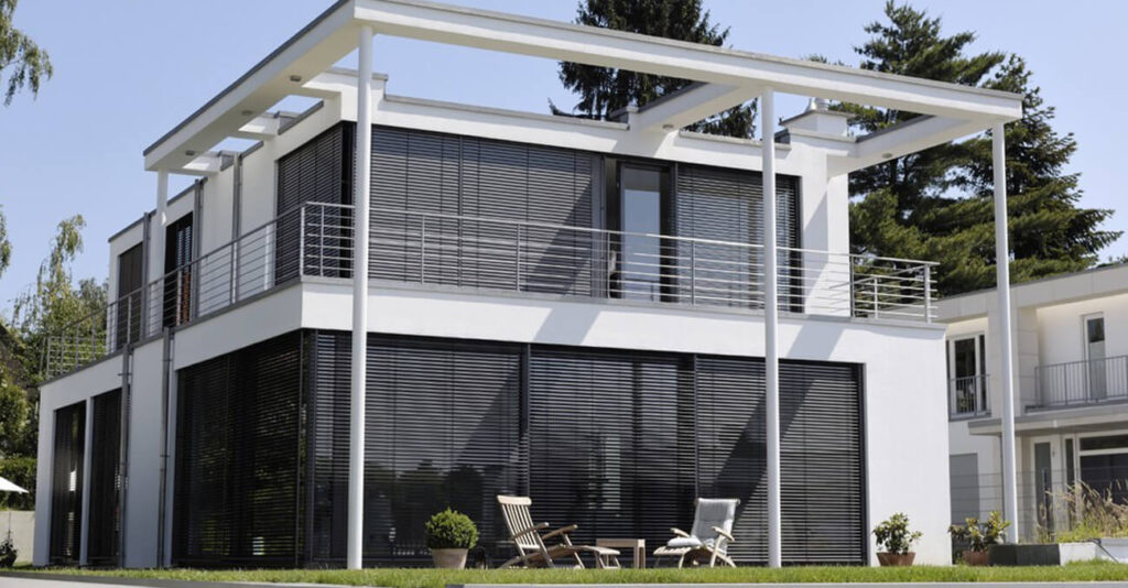 Modernes zweistöckiges Haus mit großen Fenstern und Metalljalousien, umgeben von viel Grün. Eine weiße Pergola erstreckt sich über die Terrasse, auf der zwei Holzliegestühle und kleine Topfpflanzen auf dem Rasen stehen.