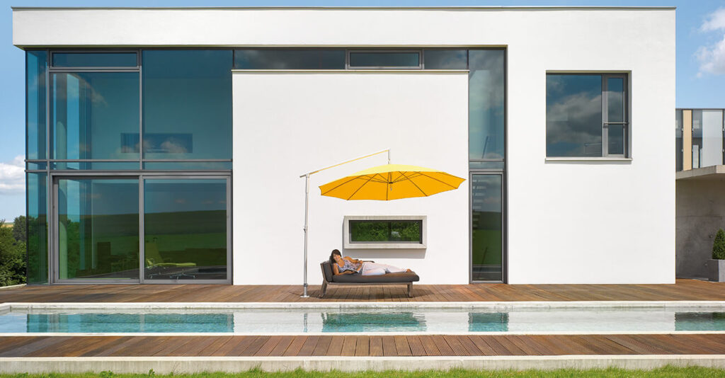 Modernes weißes Haus mit großen Fenstern, Holzterrasse und schmalem Pool. Eine Person entspannt auf einer Liege unter einem gelben Ampelschirm mit Blick auf den Pool. Die Umgebung ist hell und ruhig, der Himmel ist klar und blau.