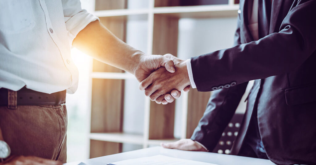 Zwei Personen geben sich in einem Büro die Hand. Eine Person trägt ein weißes Hemd und eine braune Hose, die andere einen dunklen Anzug. Sonnenlicht fällt durch ein Fenster und erhellt die Szene. Ein Tisch mit Papieren ist teilweise sichtbar.