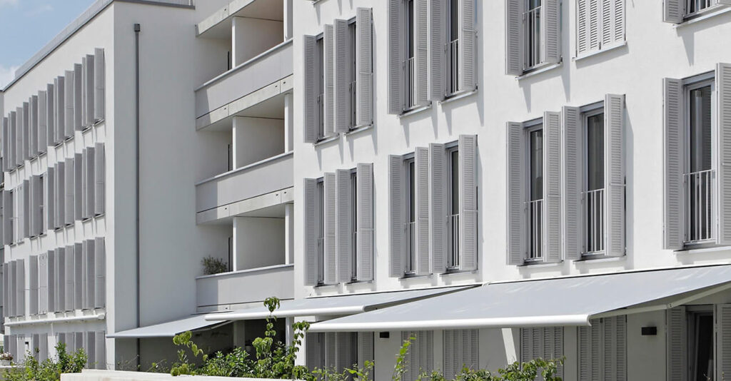Moderne Wohnhäuser mit weißer Fassade und zahlreichen Fenstern mit Faltschiebeläden. Balkone und Grünpflanzen sind sichtbar, und ausgefahrene Markisen spenden Schatten. Die Architektur ist klar und minimalistisch.