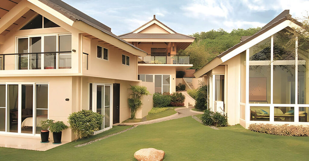 Ein moderner Wohnkomplex mit hellbraunen Wänden, großen Fenstern und schrägen Dächern ist von einem gepflegten grünen Rasen umgeben. Wege verbinden die Gebäude, und ein Felsbrocken liegt auf dem Rasen. Im Hintergrund sind Bäume unter einem bewölkten Himmel zu sehen.