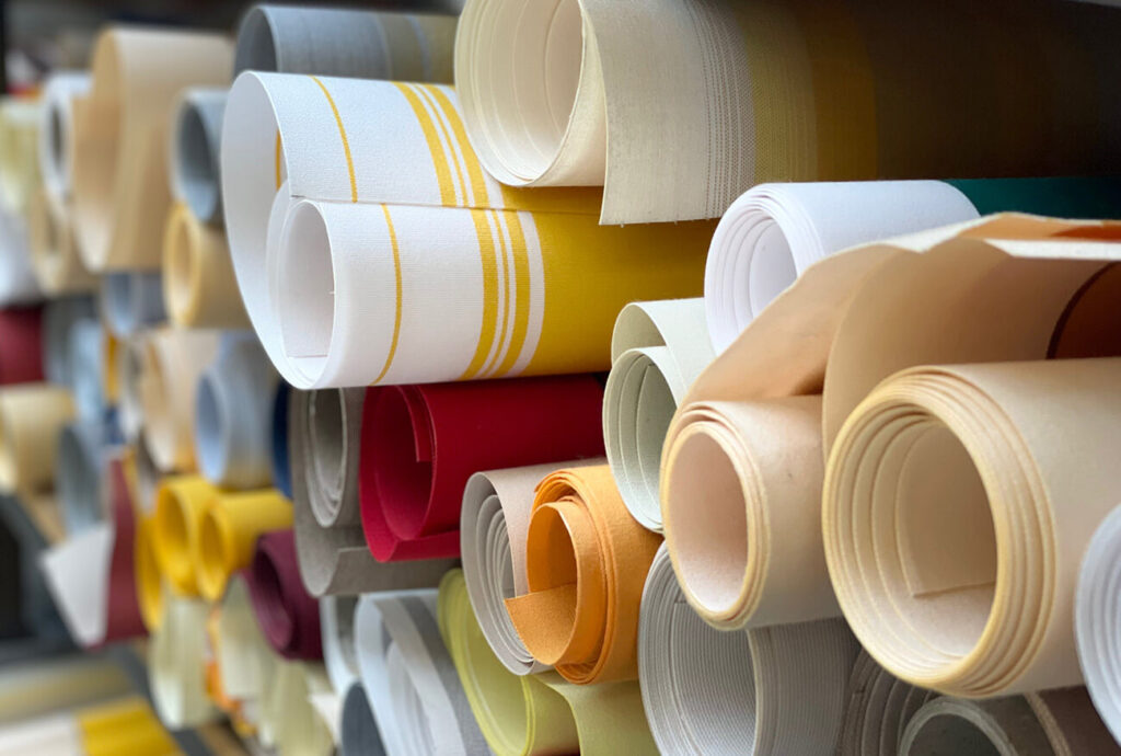 Nahaufnahme mehrerer Stoffrollen in verschiedenen Farben, darunter Gelb, Rot, Beige und Grau, die horizontal auf Regalen angeordnet sind. Einige Stoffe sind gestreift, andere einfarbig, wodurch eine optisch ansprechende und strukturierte Präsentation entsteht.