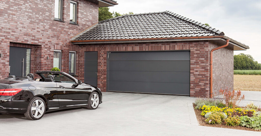 Ein modernes Backsteinhaus mit Ziegeldach und großem, geschlossenem Garagentor. In der Einfahrt parkt ein schwarzes Cabrio. Der Boden ist mit hellgrauen Fliesen gepflastert, rechts ist ein kleiner angelegter Garten zu sehen.