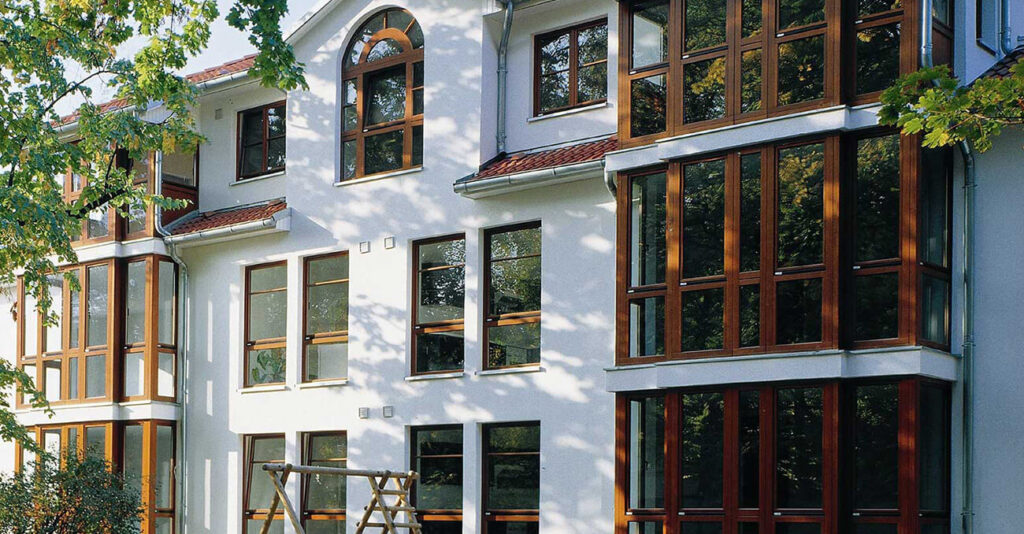 Modernes weißes Gebäude mit großen, braungerahmten Fenstern und einem schrägen Dach. Sonnenlicht fällt durch die umliegenden Bäume und wirft Schatten auf die Fassade. Im Vorgarten steht eine hölzerne Gartenschaukel. Grüne Blätter rahmen das Bild ein.