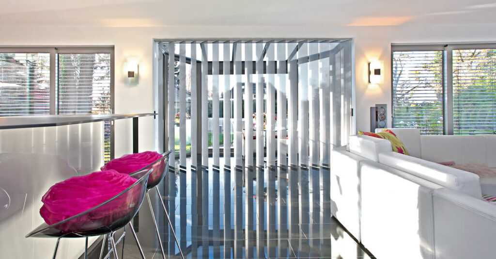 Ein modernes Wohnzimmer mit glänzend schwarzem Boden, weißen Sofas und einer Reihe vertikaler Jalousien, die zu einem verglasten Bereich führen. Barhocker mit rosa Kissen stehen links an einer weißen Theke. Große Fenster lassen viel Tageslicht herein.