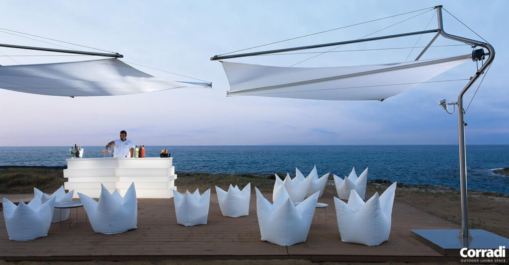 Eine Strandbar-Szene bei Sonnenuntergang. Ein Barkeeper steht hinter einer eleganten weißen Bar auf einer Holzterrasse. Weiße Sitzsäcke stehen davor, große, moderne weiße Sonnenschirme spannen sich darüber. Im Hintergrund sind das Meer und ein leicht bewölkter Himmel zu sehen.