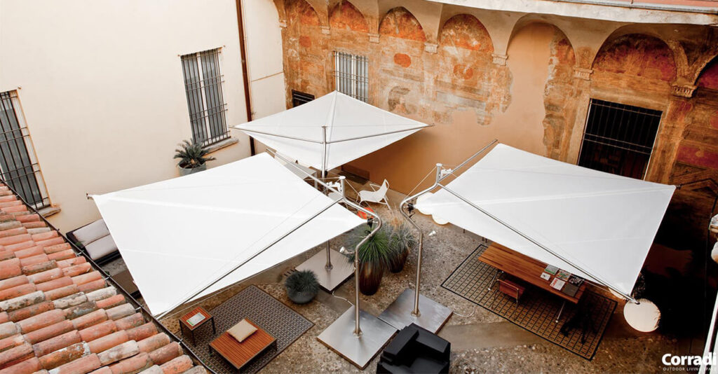 Luftaufnahme eines charmanten Innenhofs mit drei großen, weißen, quadratischen Sonnenschirmen, die Schatten spenden. Der Raum ist mit Topfpflanzen, Sitzgelegenheiten und Tischen dekoriert. Die umgebenden Wände wirken rustikal und steinern, der Boden ist mit Kopfsteinpflaster ausgelegt.