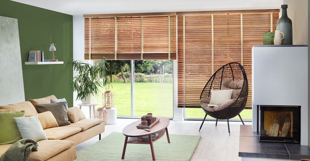 Modernes Wohnzimmer mit grüner Akzentwand, beigefarbenem Sofa mit verschiedenen Kissen und einem Couchtisch aus Holz auf einem grünen Teppich. Große Fenster mit Holzjalousien geben den Blick auf den Garten frei. Ein Hängesessel und Pflanzen sorgen für eine natürliche Note. Rechts befindet sich ein Kamin.