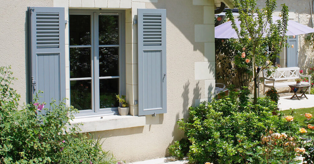 Ein charmantes Cottage mit hellblauen Klappläden und einem Fenster, umgeben von üppigem Grün und bunten Blumen. Im Hintergrund sind eine weiße Bank und ein Sonnenschirm auf einer Steinterrasse zu sehen.