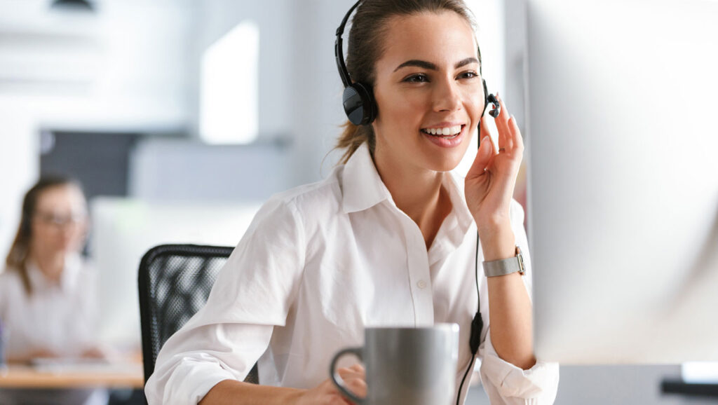 Eine lächelnde Frau mit Headset arbeitet an einem Computer in einem Büro. Sie trägt ein weißes Hemd und scheint in ein Telefonat verwickelt zu sein. Auf ihrem Schreibtisch steht eine graue Tasse. Im Hintergrund ist ein verschwommener Kollege zu sehen.