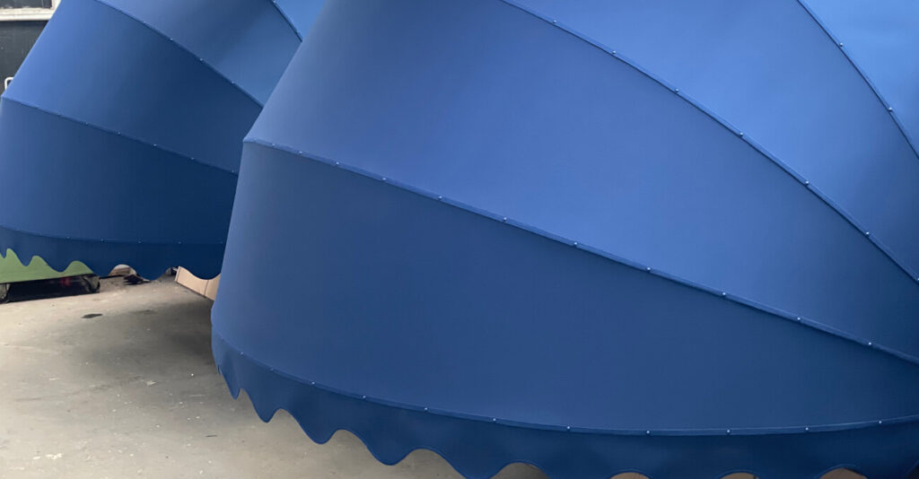 Nahaufnahme von zwei großen, überlappenden blauen Markisen mit gewellten Kanten. Der Stoff hat eine glatte Textur mit sichtbaren Nähten. Der Boden darunter ist eine schlichte Betonoberfläche.