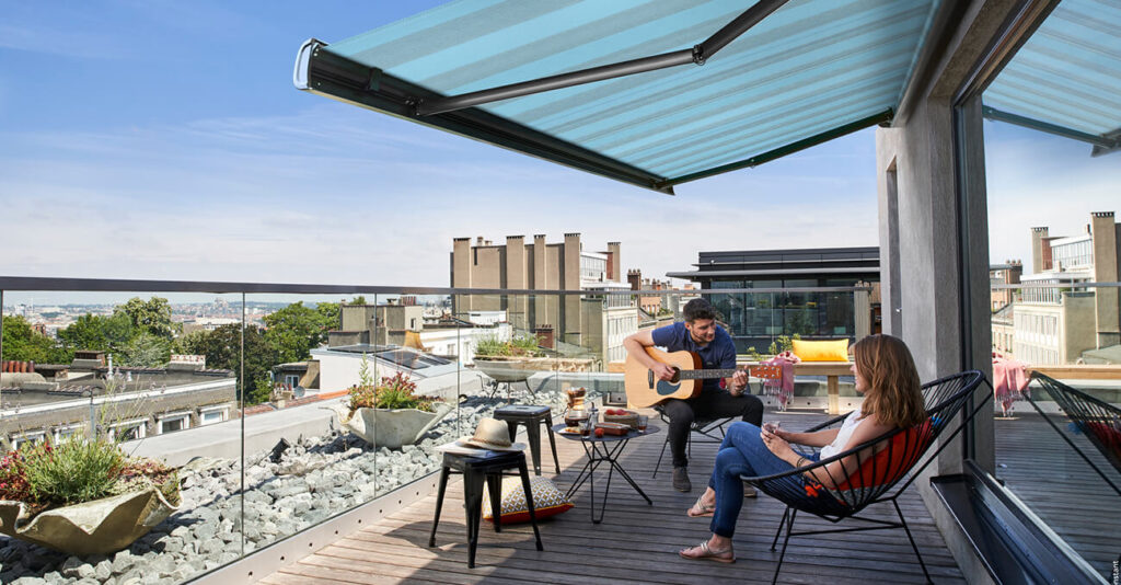 Zwei Personen entspannen auf einer Dachterrasse. Einer spielt Gitarre, der andere sitzt da und hört zu. Die Terrasse besticht durch eine blaue Markise, Topfpflanzen und einen Blick auf die Stadtgebäude unter klarem Himmel. Auf einem kleinen Tisch stehen Getränke und Snacks bereit.