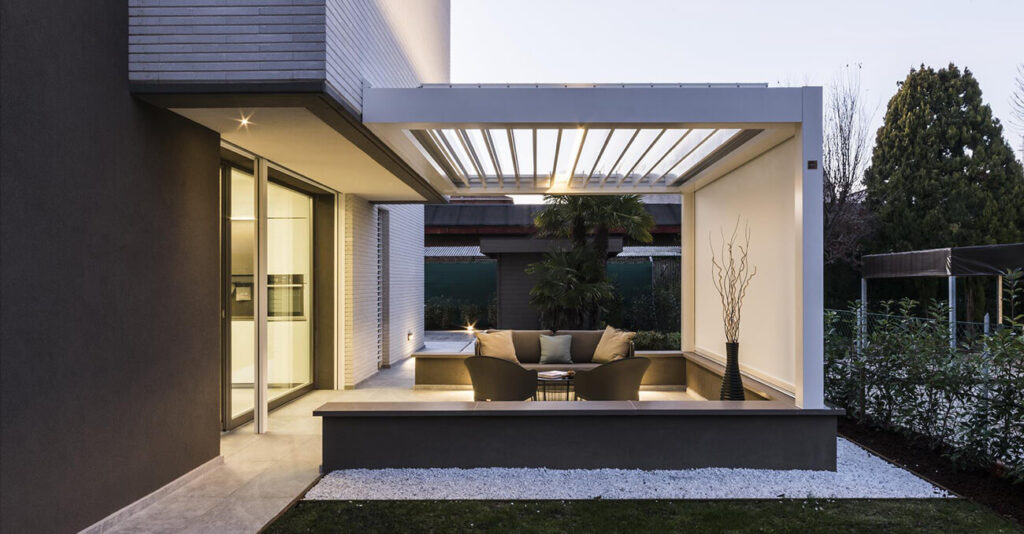 Moderne Außenterrasse mit eleganter Pergola, beigen Sofas und einem Couchtisch aus Glas. Der Bereich ist von Grünflächen und minimalistischer Architektur umgeben. Sanfte Beleuchtung sorgt für eine gemütliche Atmosphäre.
