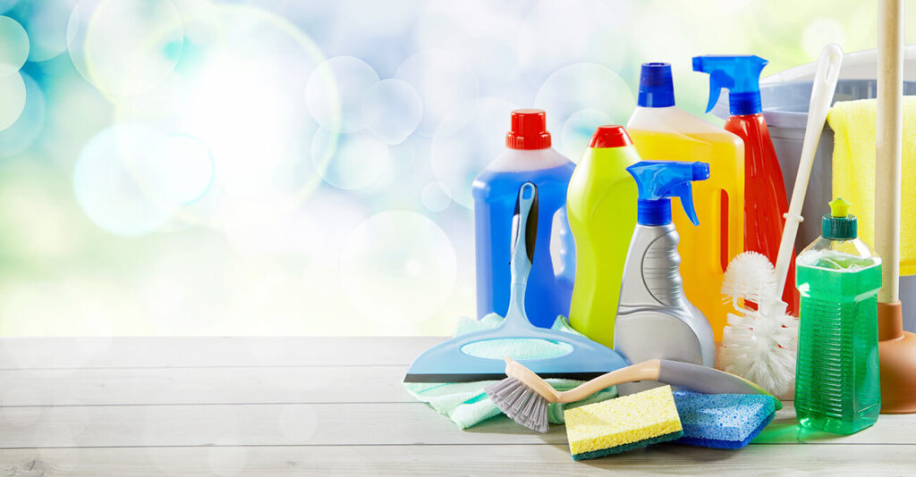 Auf einer Holzfläche vor unscharfem Hintergrund sind verschiedene bunte Reinigungsmittel arrangiert. Zu den Artikeln gehören Sprühflaschen, Schwämme, Bürsten und Reinigungsmittelbehälter, die Werkzeuge für die Haushaltsreinigung präsentieren.