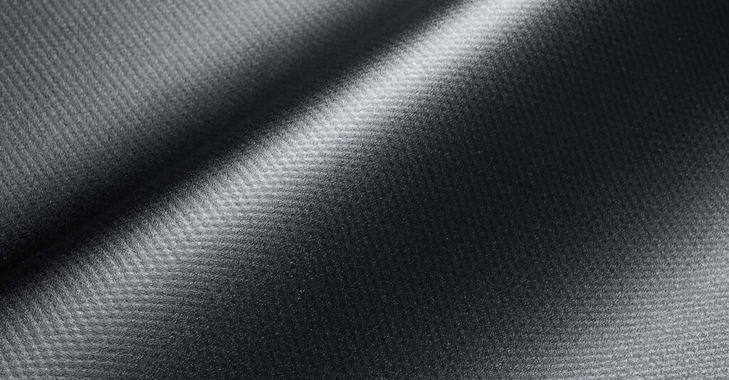 Nahaufnahme eines glänzenden, strukturierten schwarzen Stoffes mit diagonal verlaufenden Linien. Die Beleuchtung erzeugt einen subtilen Farbverlauf, der die glatte Oberfläche und das komplizierte Webmuster hervorhebt.