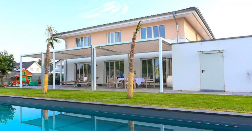 Modernes zweistöckiges Haus mit großen Fenstern, weißer Fassade und Flachdach. Der Hinterhof verfügt über einen Swimmingpool, eine Terrasse mit Essbereich, Liegestühlen und Palmen. Links ist ein Kinderspielhaus zu sehen.