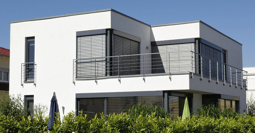 Modernes zweistöckiges weißes Haus mit Flachdach und großen Fenstern, teilweise mit Raffstoren. Ein Balkon mit Metallgeländer umschließt das Obergeschoss. Im Vordergrund sind grüne Büsche und ein geschlossener Sonnenschirm unter einem strahlend blauen Himmel zu sehen.