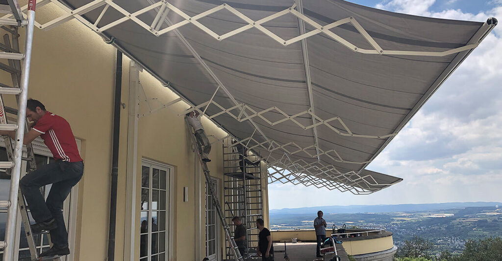 Monteure installieren große Markisen an der Außenseite eines Gebäudes. Eine Person steht auf einer Leiter, andere arbeiten am Boden. Der Blick hinter ihnen zeigt eine weite Landschaft mit Hügeln unter einem teilweise bewölkten Himmel.