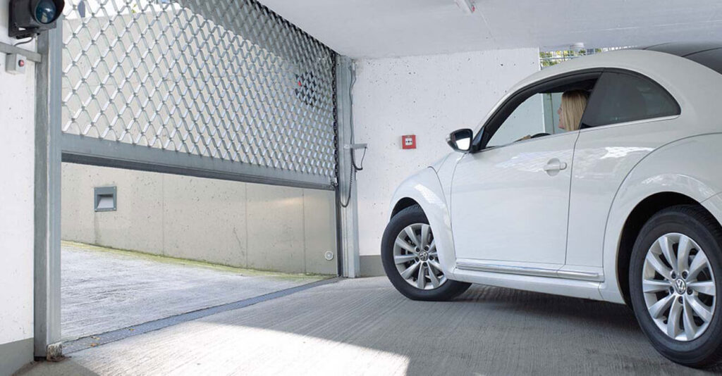 Ein weißes Auto fährt aus einer Garage mit Metalltor. Der Fahrer, eine Person mit langen blonden Haaren, ist durch das Fenster sichtbar. Die Garage hat Betonboden und -wände. Rechts an der Wand befindet sich ein roter Knopf.