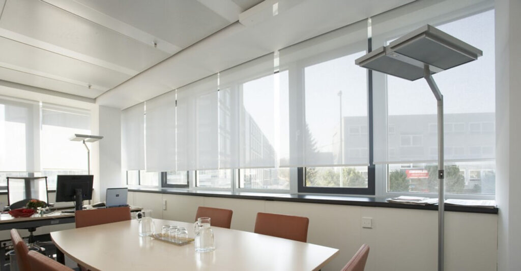 Ein moderner Konferenzraum mit einem Tisch und vier Stühlen. Große Fenster mit lichtdurchlässigen Rollos sorgen für Tageslicht. Im Hintergrund stehen ein Computer und Büromaterial auf einem Schreibtisch, daneben steht eine Stehlampe.