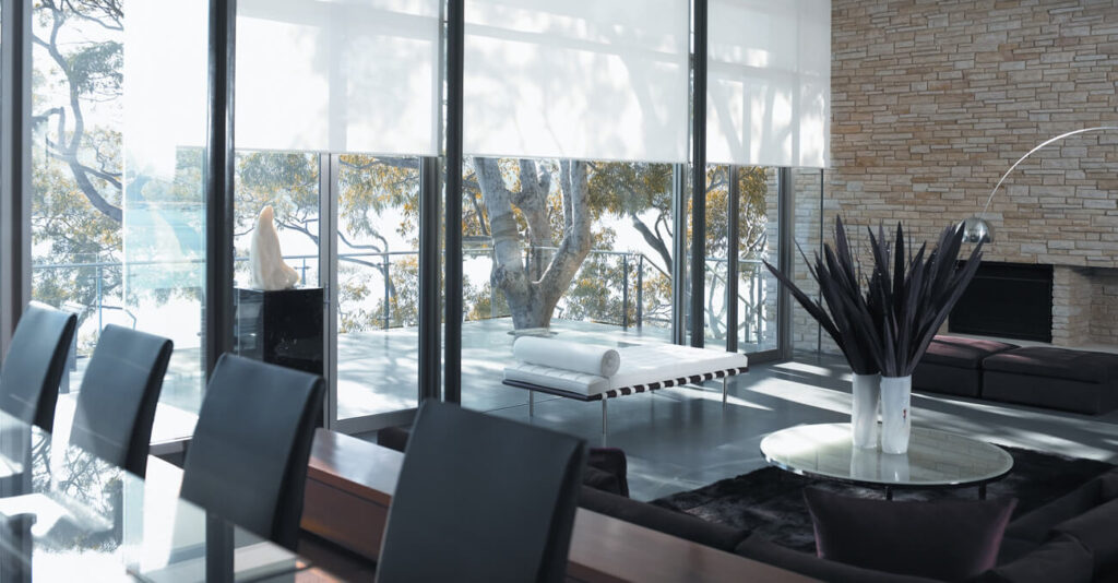 Modernes Wohnzimmer mit großen Fenstern und Blick auf Bäume. Zur Ausstattung gehören schwarze Stühle, ein minimalistisches Sofa, eine Steinwand mit Kamin und ein runder Tisch mit hohen Pflanzen. Natürliches Licht durchflutet den Raum und schafft eine luftige Atmosphäre.