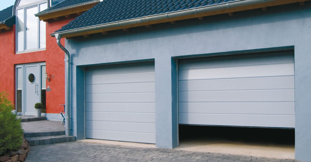 Ein modernes Haus mit roter Fassade verfügt über zwei nebeneinanderliegende graue Garagentore. Ein Tor ist teilweise geöffnet, das andere geschlossen. Die Auffahrt ist mit Steinfliesen gepflastert, und eine kleine Treppe führt zur Haustür.