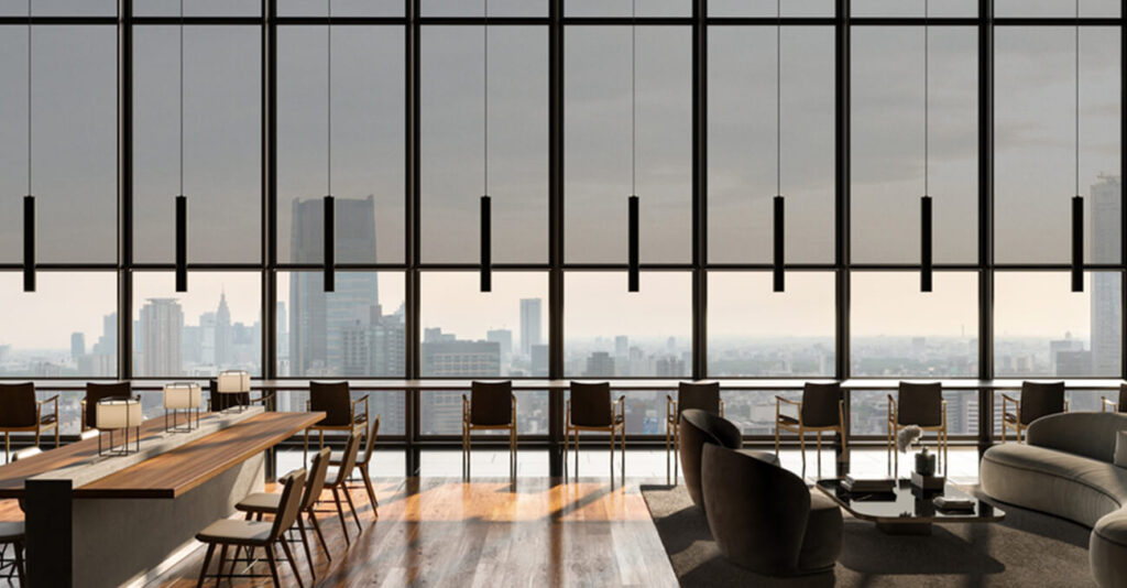 Eine moderne, geräumige Lounge mit raumhohen Fenstern bietet einen Panoramablick auf die Skyline der Stadt. Der Raum verfügt über Holztische, bequeme Sitzgelegenheiten und Hängelampen, die eine elegante, elegante Atmosphäre schaffen.