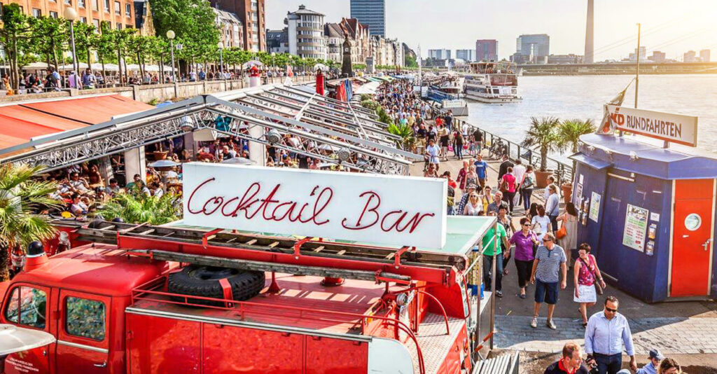 Eine lebhafte Szene am Flussufer zeigt eine belebte Promenade mit Menschen, die unter rot-weißen Markisen spazieren gehen und in Cafés sitzen. Auf einem roten Lastwagen prangt ein „Cocktailbar“-Schild. Im Hintergrund sind der Fluss, Boote und die Skyline der Stadt zu sehen.