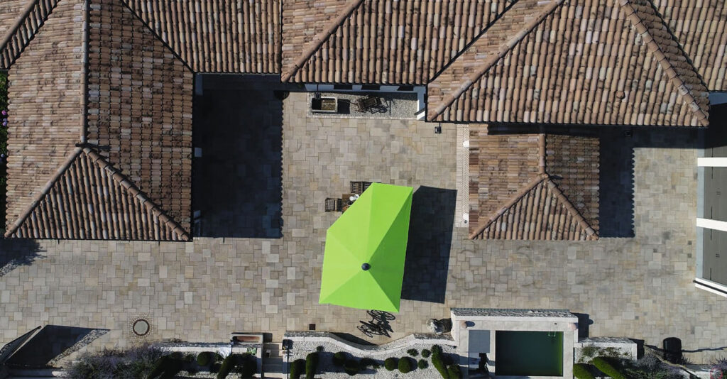 Luftaufnahme eines Innenhofs mit einem großen grünen Sonderschirm auf beigen Steinfliesen. Die umliegenden Gebäude haben braune Ziegeldächer. Am unteren Bildrand sind gepflegte Büsche und ein kleiner Gartenbereich zu sehen.