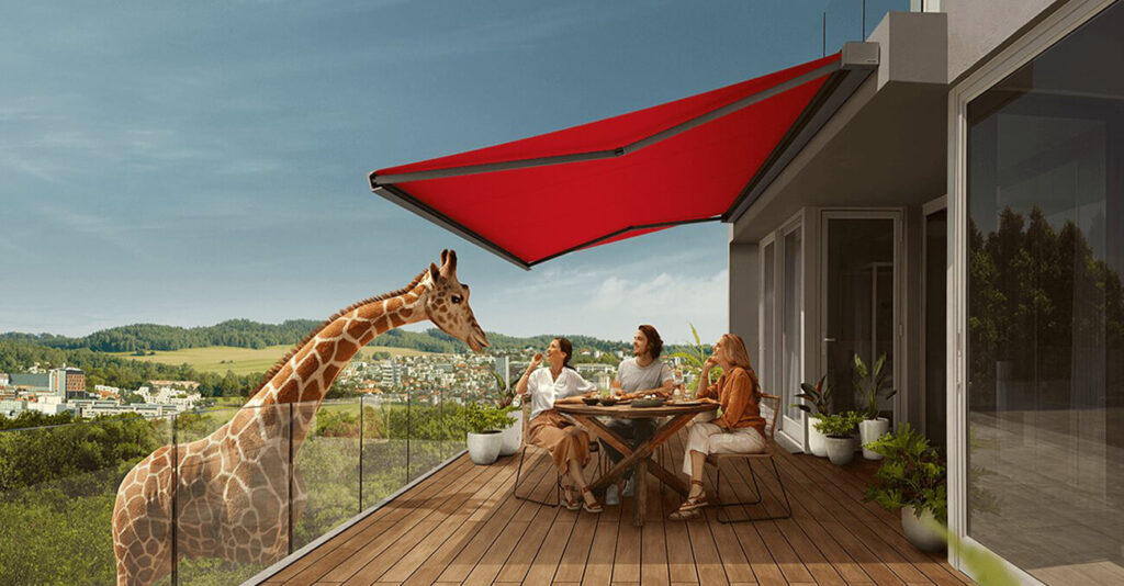 Drei Personen sitzen an einem Terrassentisch unter einer großen roten Markise und genießen ihr Essen. Eine Giraffe steht in der Nähe und beobachtet sie neugierig. Im Hintergrund ist eine malerische Aussicht auf grünes Grün und entfernte Gebäude zu sehen.