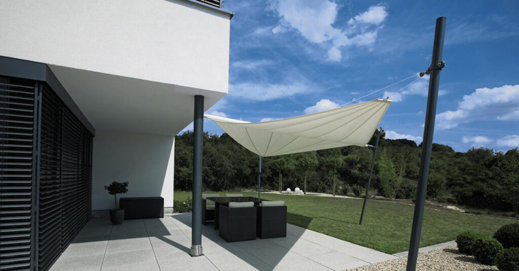 Eine moderne Terrasse mit einem weißen Sonnensegel spendet Schatten über einem Sitzbereich mit dunklen Korbmöbeln. Die Terrasse grenzt an ein weißes Gebäude mit großen, dunklen Fensterläden. Im Hintergrund sind ein grüner Rasen und Bäume unter einem blauen Himmel mit Wolken zu sehen.