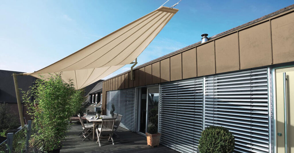 Eine moderne Dachterrasse mit Sitzgelegenheiten, Pflanzen und einem beigen dreieckigen Sonnensegel. Die Außenfassade besticht durch große Fenster mit Metalljalousien. Der klare, blaue Himmel sorgt für eine helle Atmosphäre.