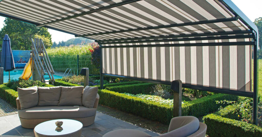 Eine moderne Außenterrasse mit gestreifter Terrassenmarkise. Darunter stehen gepolsterte Korbstühle und ein kleiner Tisch. Ein üppiger grüner Garten umgibt den Bereich. Im Hintergrund sind ein Pool und ein Sonnenschirm zu sehen.