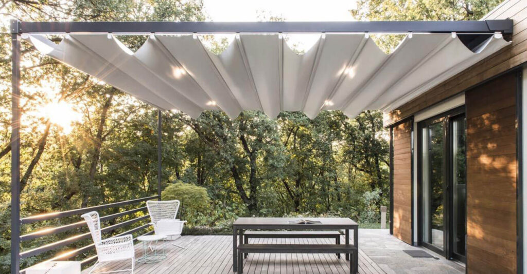 Moderne Außenterrasse mit ausfahrbarer Markise, Holzdeck und Esstisch. Umgeben von üppigen grünen Bäumen fällt Sonnenlicht durch das Laub. Zwei Stühle stehen seitlich.