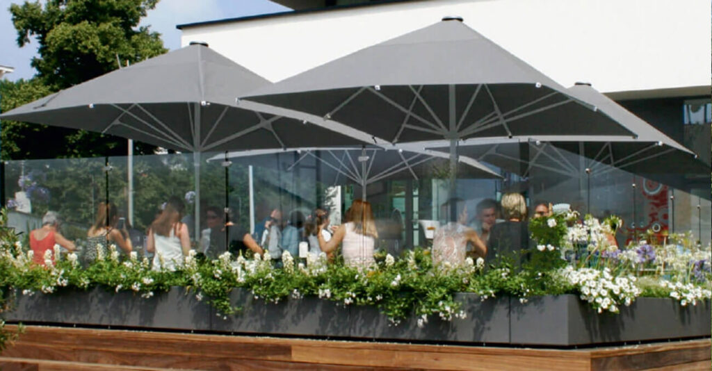 Straßencafé-Szene mit Menschen unter großen grauen Sonnenschirmen, umgeben von Glaswänden und üppigem Grün. Im Vordergrund sind Blumen zu sehen, im Hintergrund ein modernes Gebäude. Sonniges Wetter.