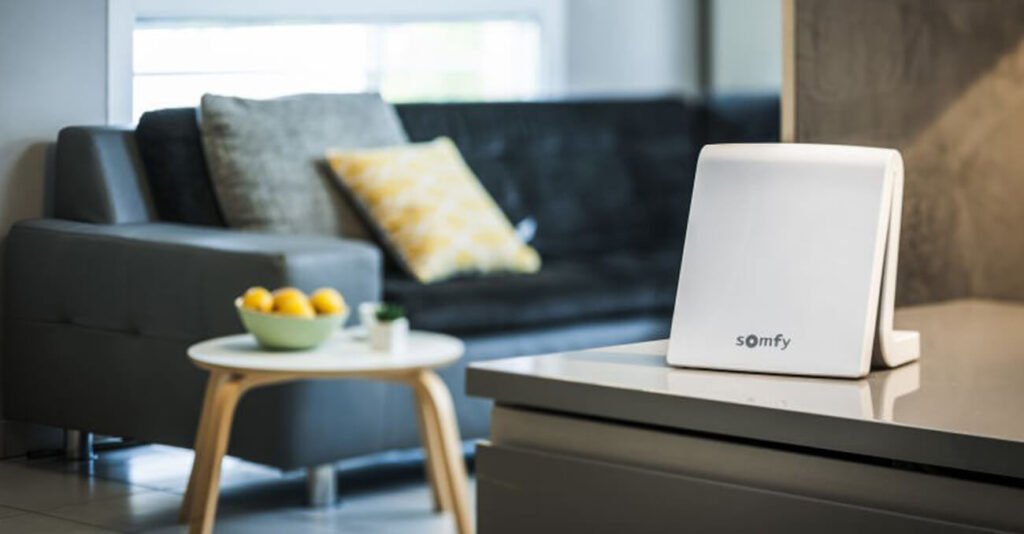 Nahaufnahme eines weißen Somfy-Geräts auf einer Küchentheke. Im Hintergrund ist ein modernes Wohnzimmer mit einem dunkelgrauen Sofa mit gelbem Kissen und einem kleinen weißen Couchtisch mit einer Schale Zitronen zu sehen. Ein Fenster sorgt für natürliches Licht.