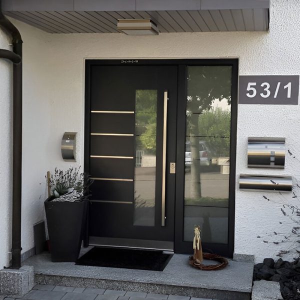 Moderne schwarze Haustür mit horizontalen Metallstreifen und Glasfüllung. Neben der Tür stehen eine Topfpflanze und drei silberne Briefkästen an einer weißen Wand. Neben der Tür prangt die Hausnummer 53/1. Ein kleines Dach überdeckt den Eingang.