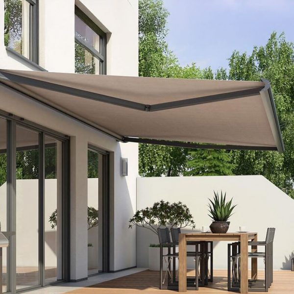 Eine moderne Terrasse mit einer einziehbaren beigen Markise an einem weißen Gebäude. Die Terrasse hat einen Holzboden, einen Esstisch mit vier Stühlen und Topfpflanzen. Große Glastüren führen hinein und im Hintergrund sind Bäume zu sehen.